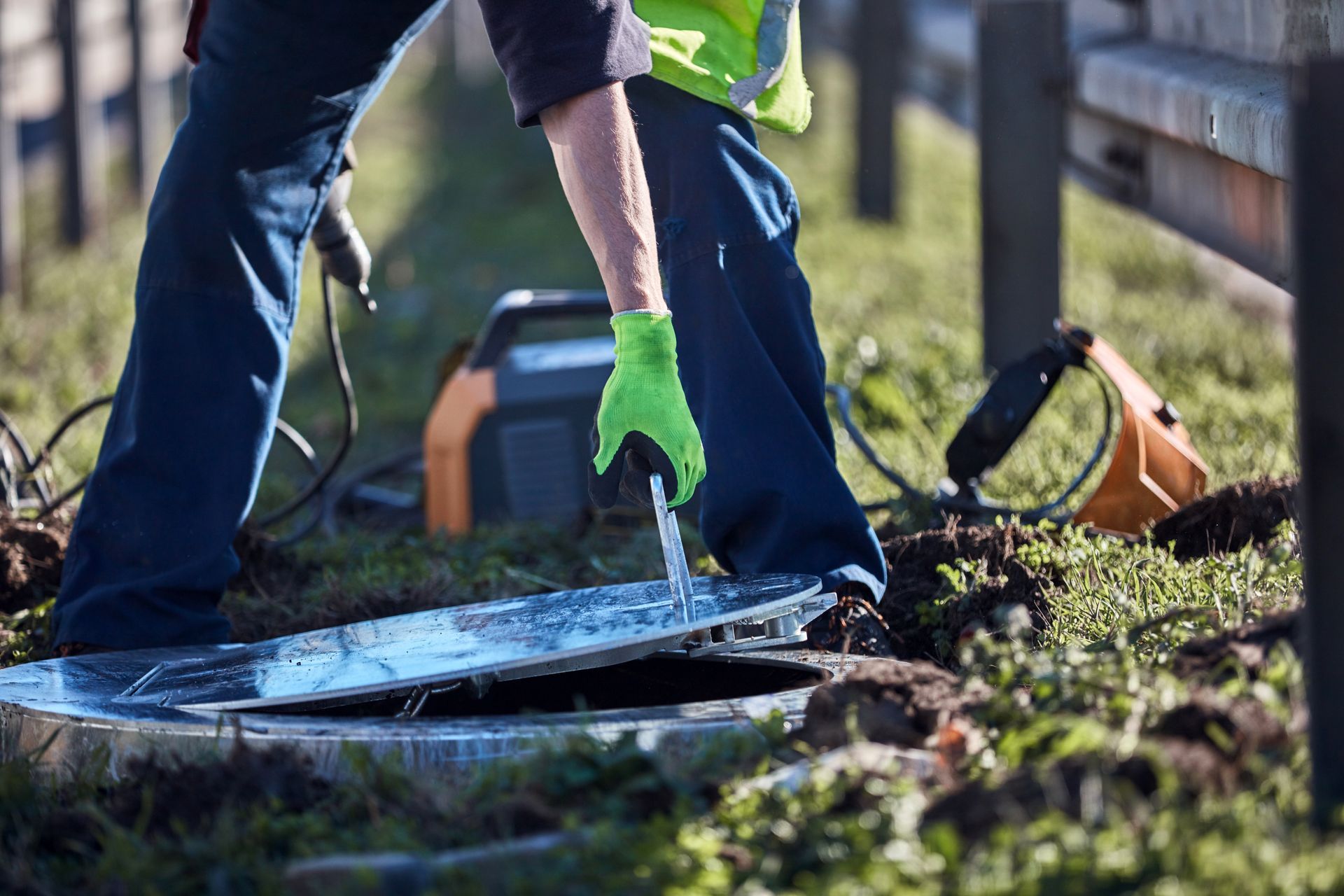 Worker opening a manhole cover outdoors, wearing gloves, reflective vest.