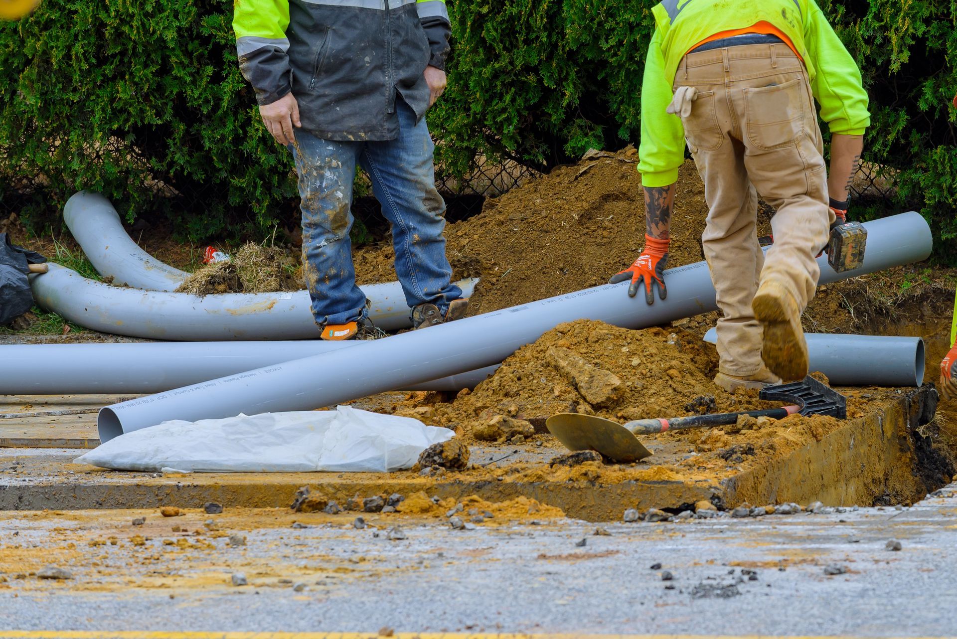Construction workers near opened trench with gray pipes and dirt. One worker is walking.
