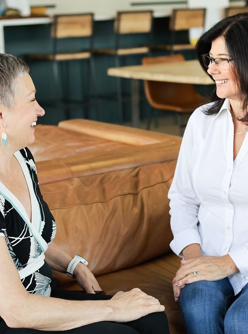 Two women smiling, seated and talking; casual interior setting with wood furniture.