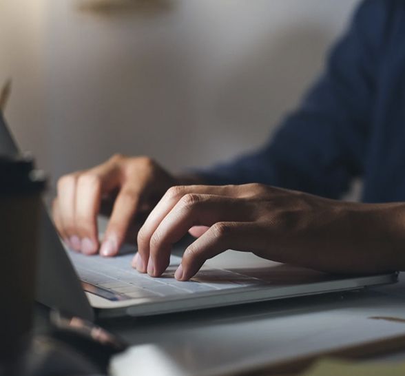Hands typing on a laptop keyboard. Dark setting, partially visible blue sleeve.