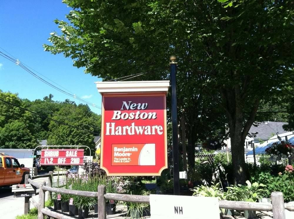 Sign for New Boston Hardware, red and white, in front of greenery and a white building.