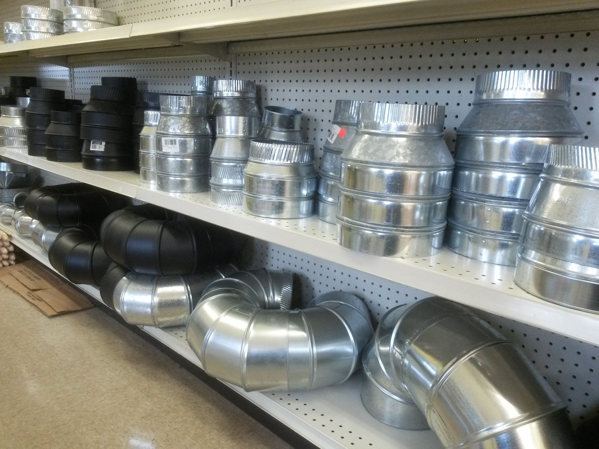 Shelves stocked with galvanized and black metal ductwork fittings in a hardware store.