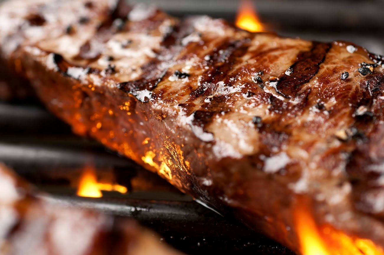 Primo piano di una bistecca alla griglia con segni di bruciatura e fiamme sottostanti.