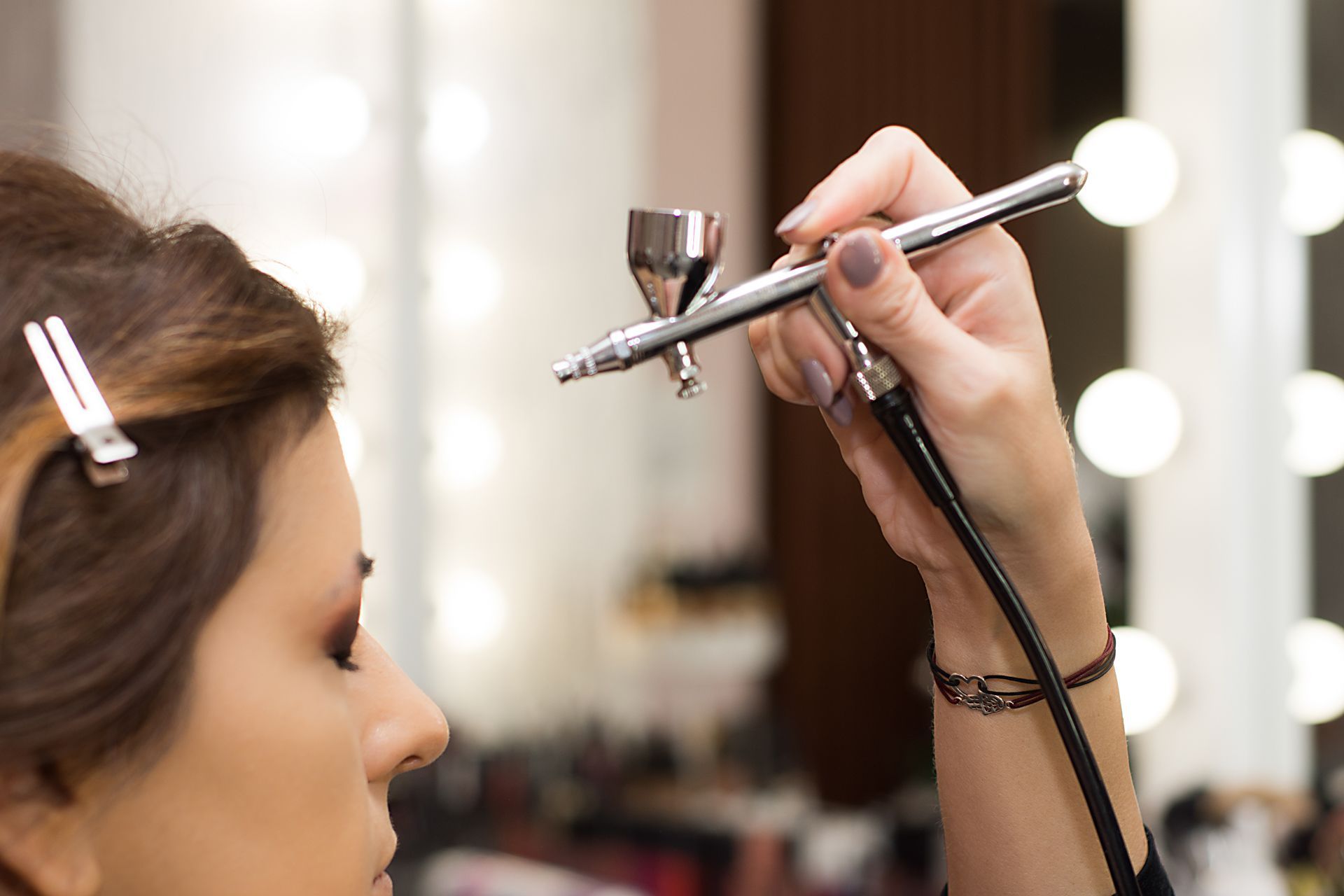 Makeup artist airbrushing face in a salon, with mirror lights in the background
