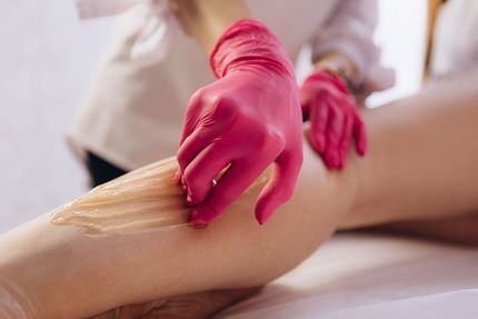 Gloved hands apply wax to a leg during a waxing treatment