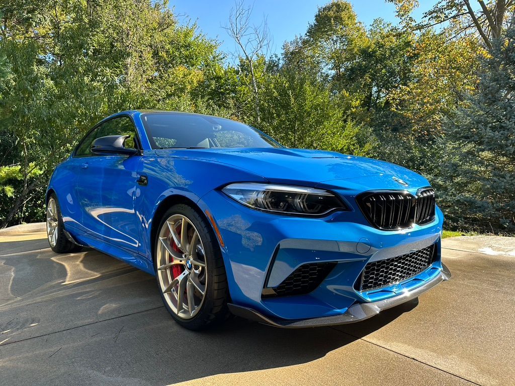 Blue BMW M2 coupe parked on a paved surface, with red brake calipers and trees in the background.