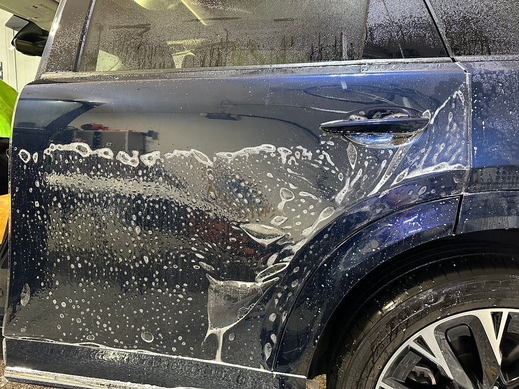 Dark blue car with soap foam; a worker is washing the vehicle.