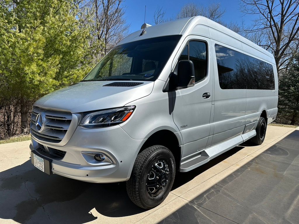 Silver Mercedes-Benz Sprinter van parked on a paved surface, black wheels, sunny day.