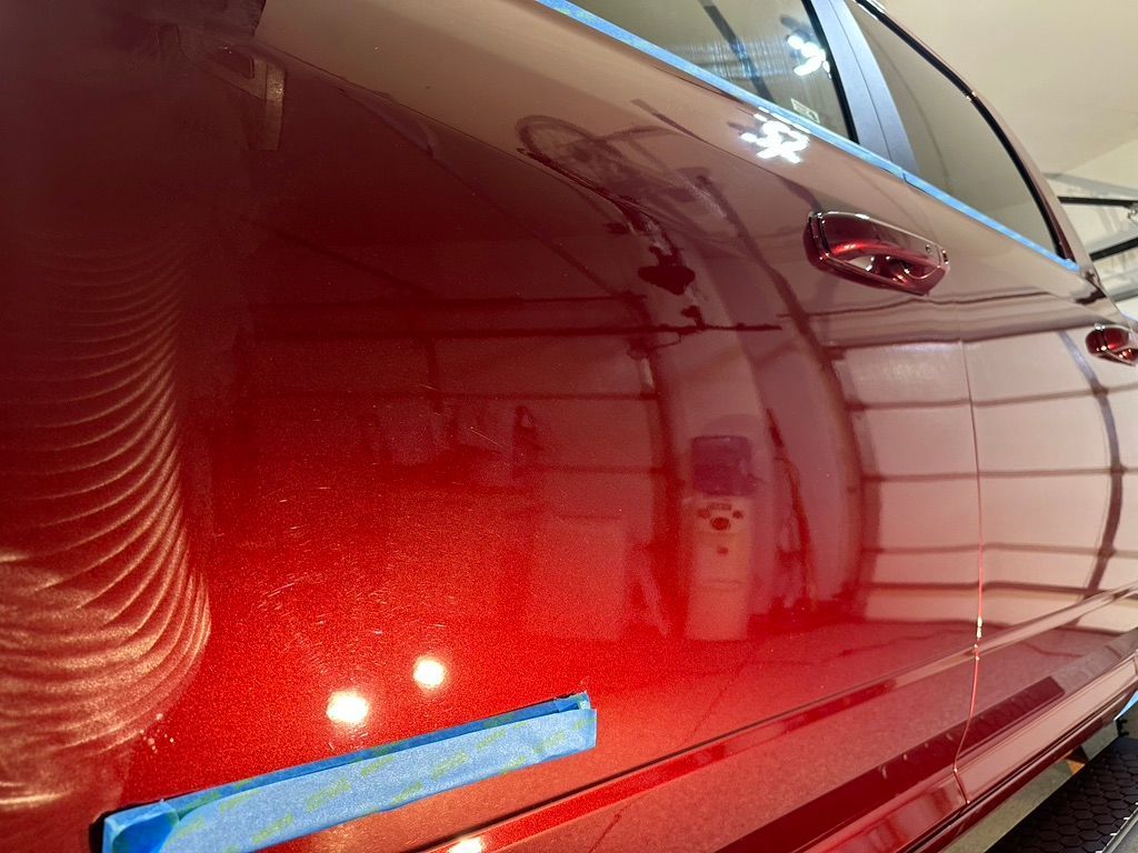 Red car door with scratches being polished; visible reflection of garage and equipment.