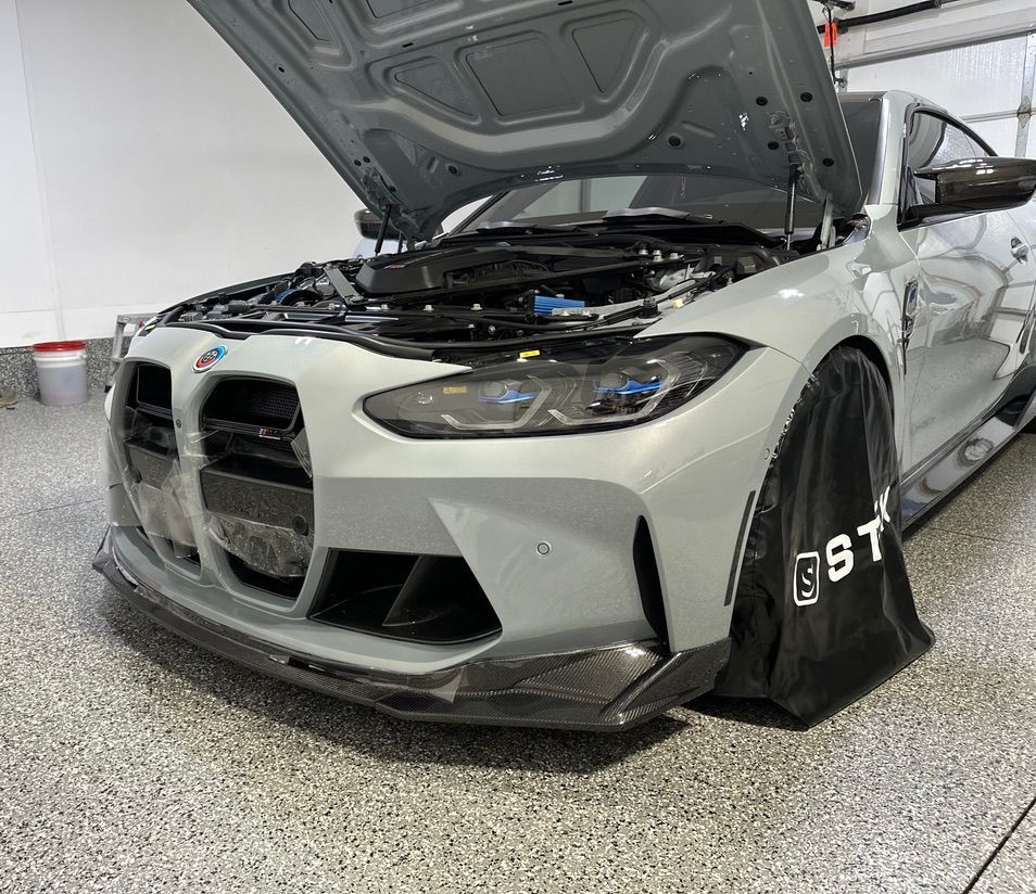 Gray BMW M4 with open hood; black front spoiler, black protective cover on the wheel, garage setting.