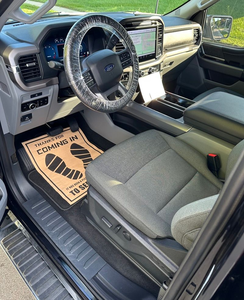 Interior view of a Ford truck. Gray seats, black dashboard, steering wheel covered in plastic. Footprint-shaped welcome mat.