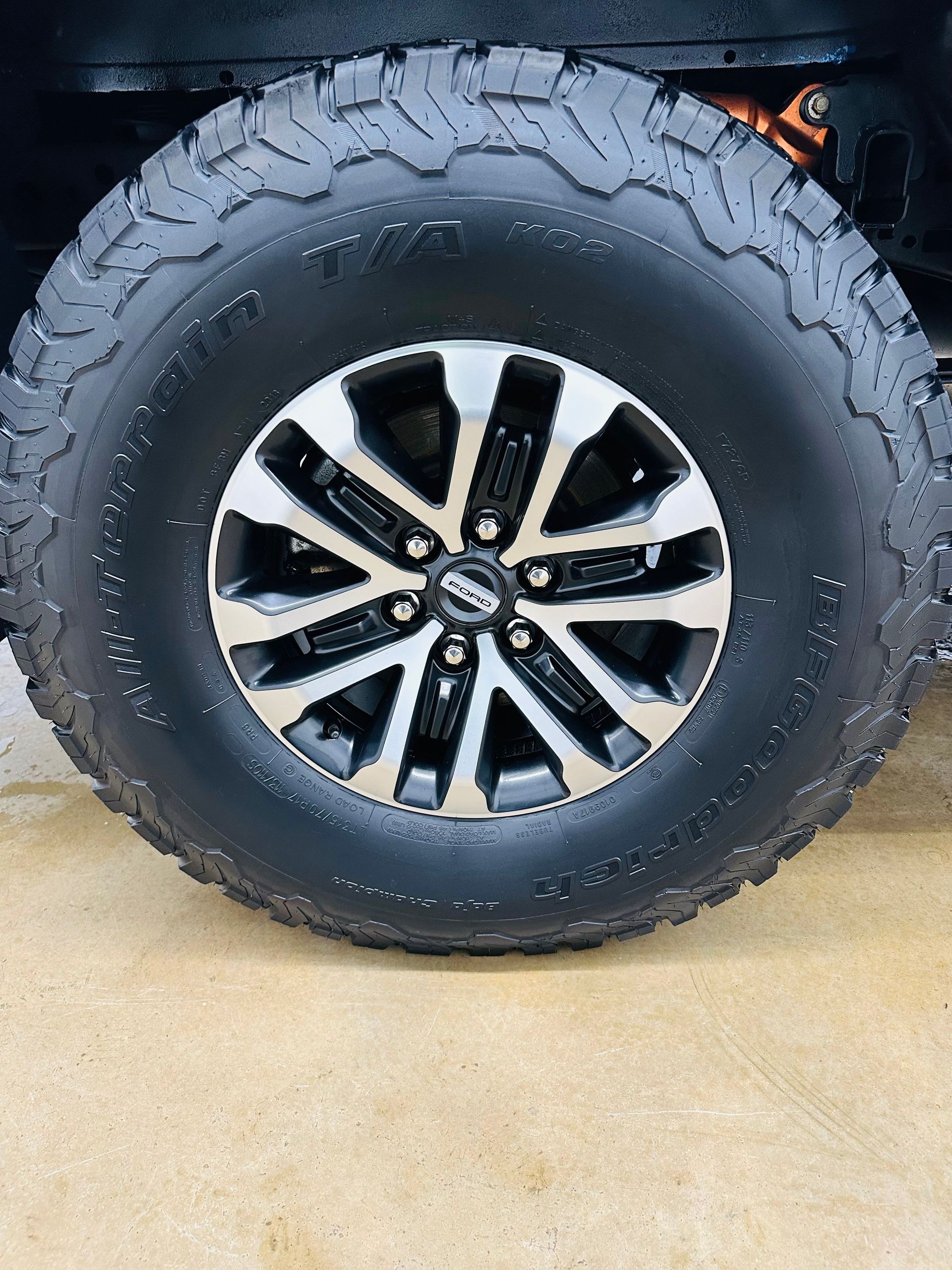Black off-road tire and silver and black rim on a vehicle, on a light-colored surface.