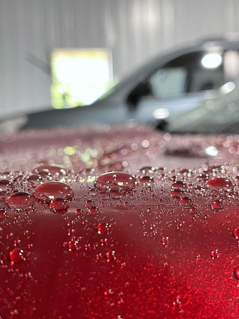 Close-up of red car hood with water droplets, partially obscuring a gray vehicle in a bright setting.