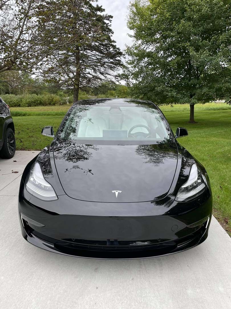 Black Tesla Model 3 parked on a concrete driveway, trees in the background, cloudy day.