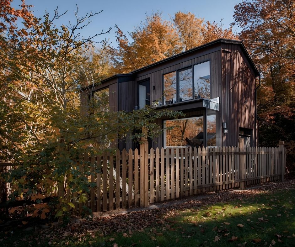 A modern two-story dark wood cabin with large windows set in an autumn forest behind a light-colored wooden picket fence.