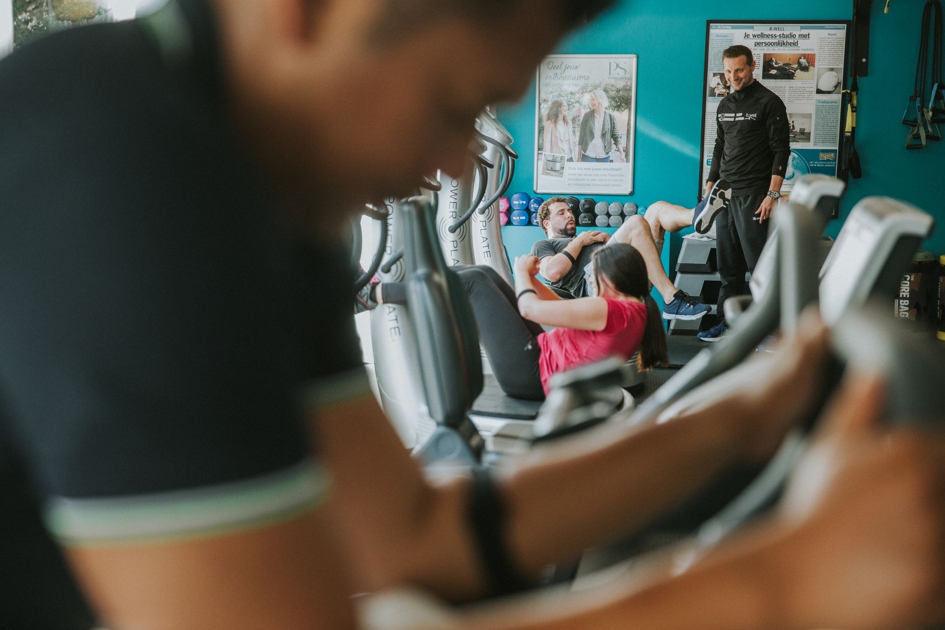 Person on a gym machine, others exercising with a trainer in a brightly lit room.