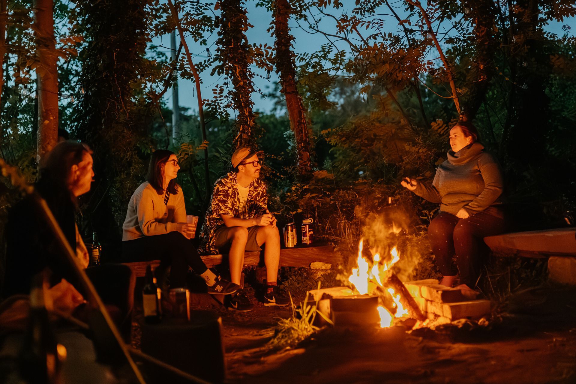 People sit around a campfire in a forest at dusk, listening to someone speaking.