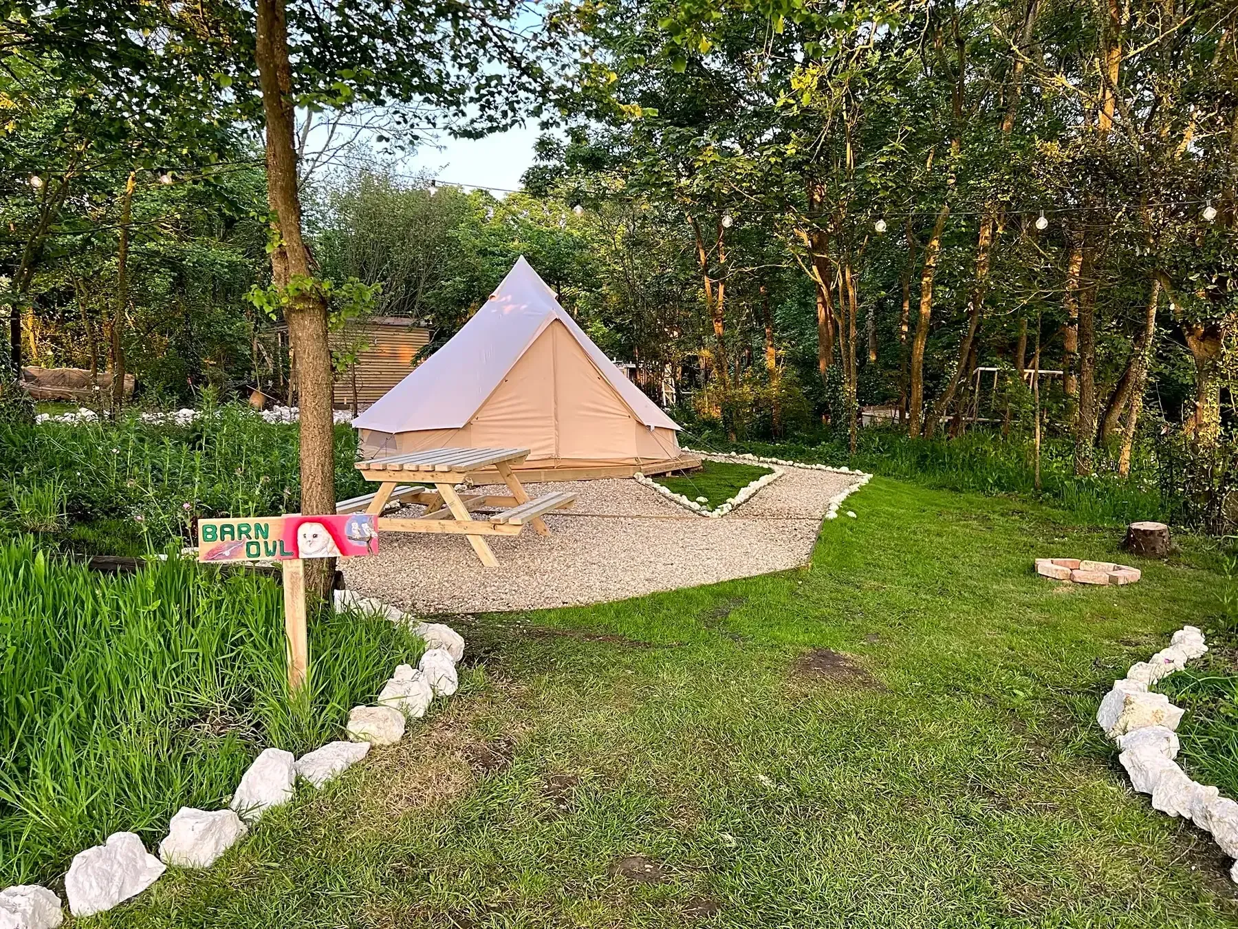 Tan canvas tent with picnic table on a gravel patch in a grassy area with trees and a pathway lined with white rocks.