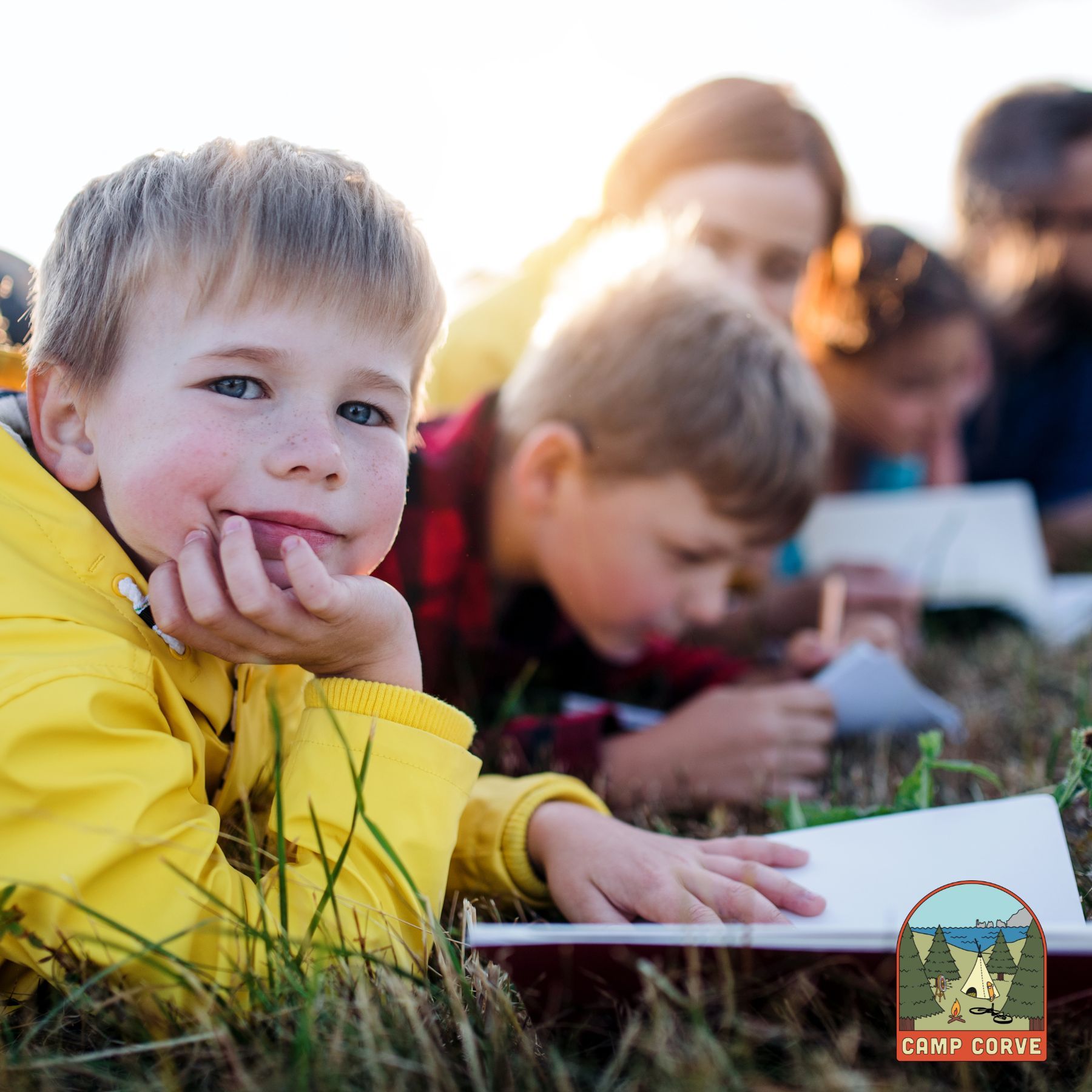 A young child in a yellow jacket smiles while lying on grass with others reading books.