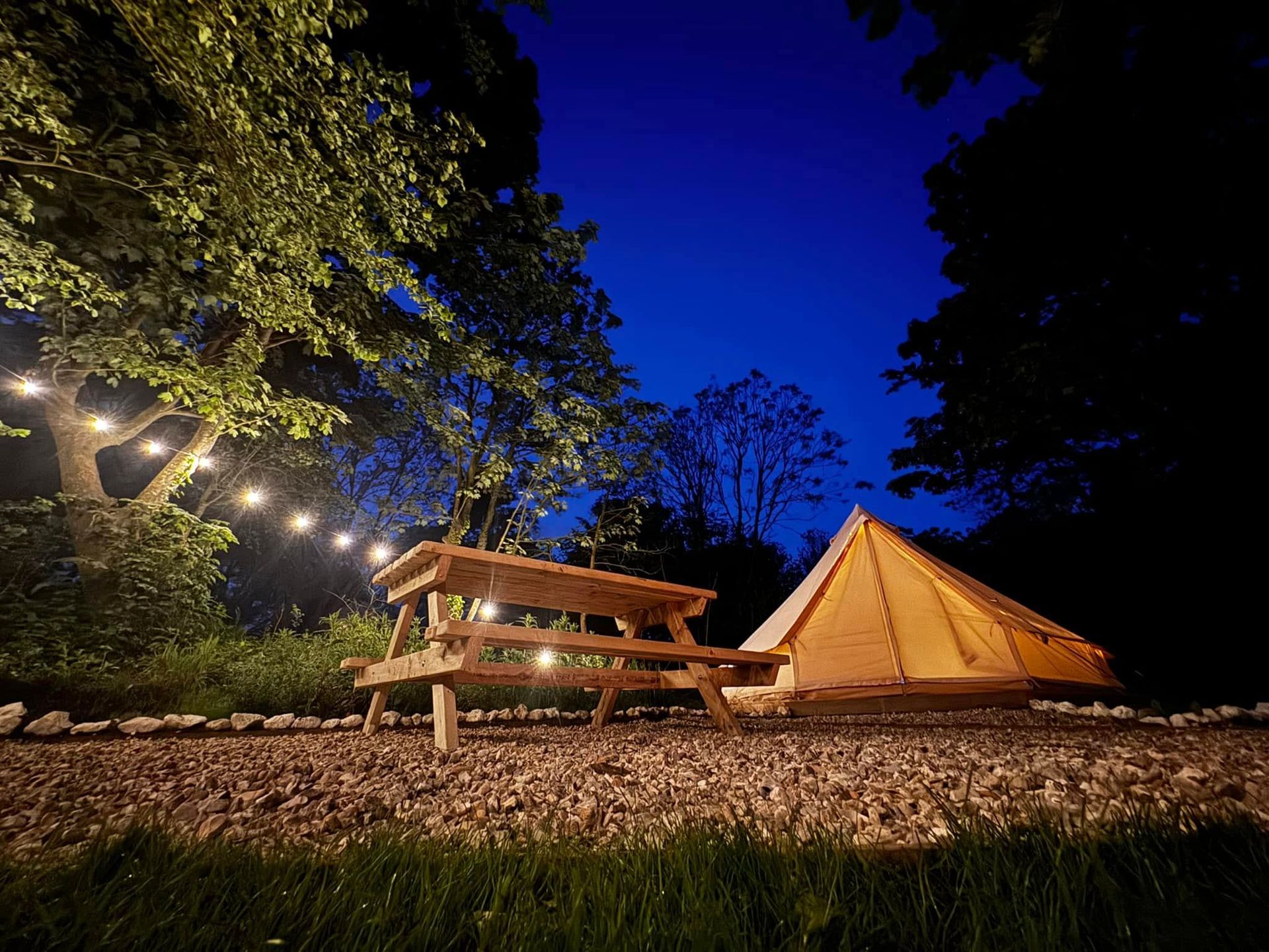 Campsite at night with picnic table, tent, and string lights, surrounded by trees under a dark blue sky.