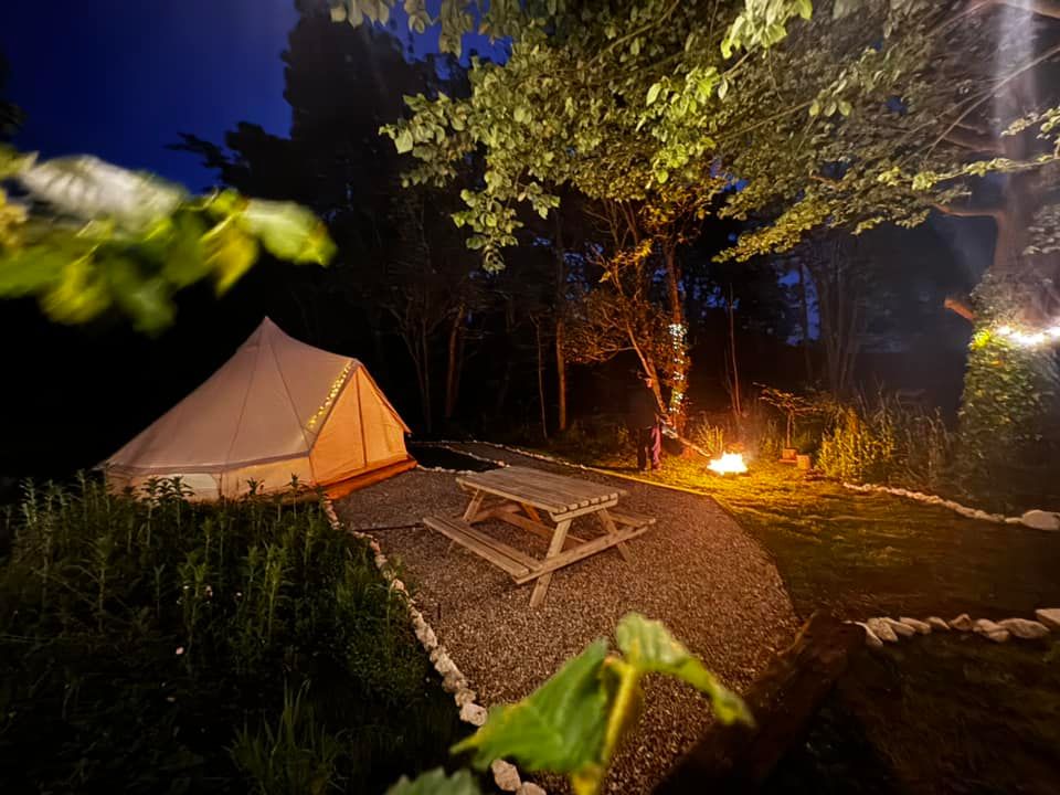 A lit canvas tent and picnic table on a gravel path at night, with fire pit and tree with fairy lights.