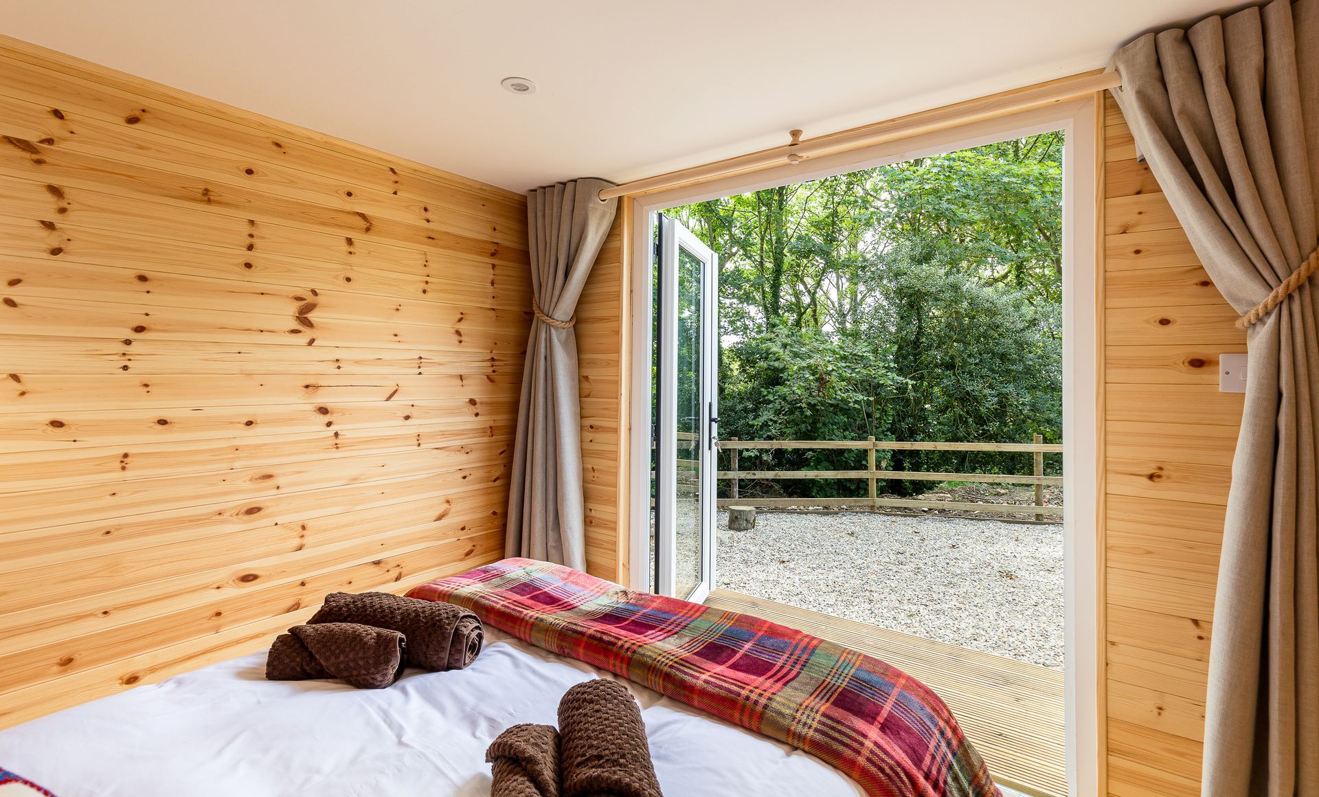 Bedroom with wooden walls, bed, and open doors to a gravel patio and trees.