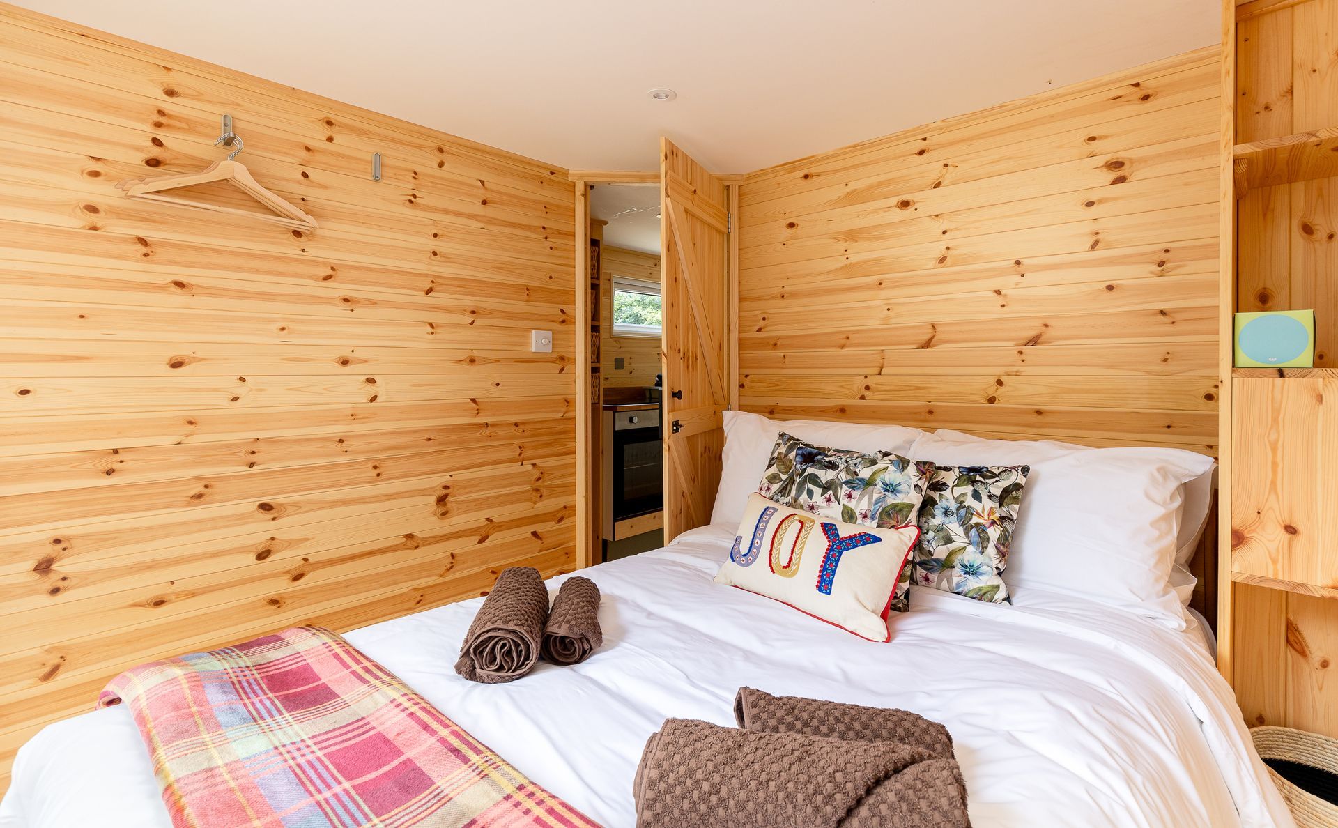 Cozy bedroom with wooden paneling, a bed with pillows and blanket, and a small kitchenette visible.