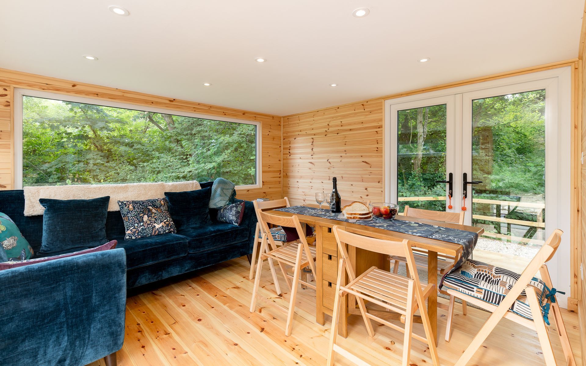 Cozy cabin interior with blue couch, wooden table and chairs, and large windows overlooking trees.