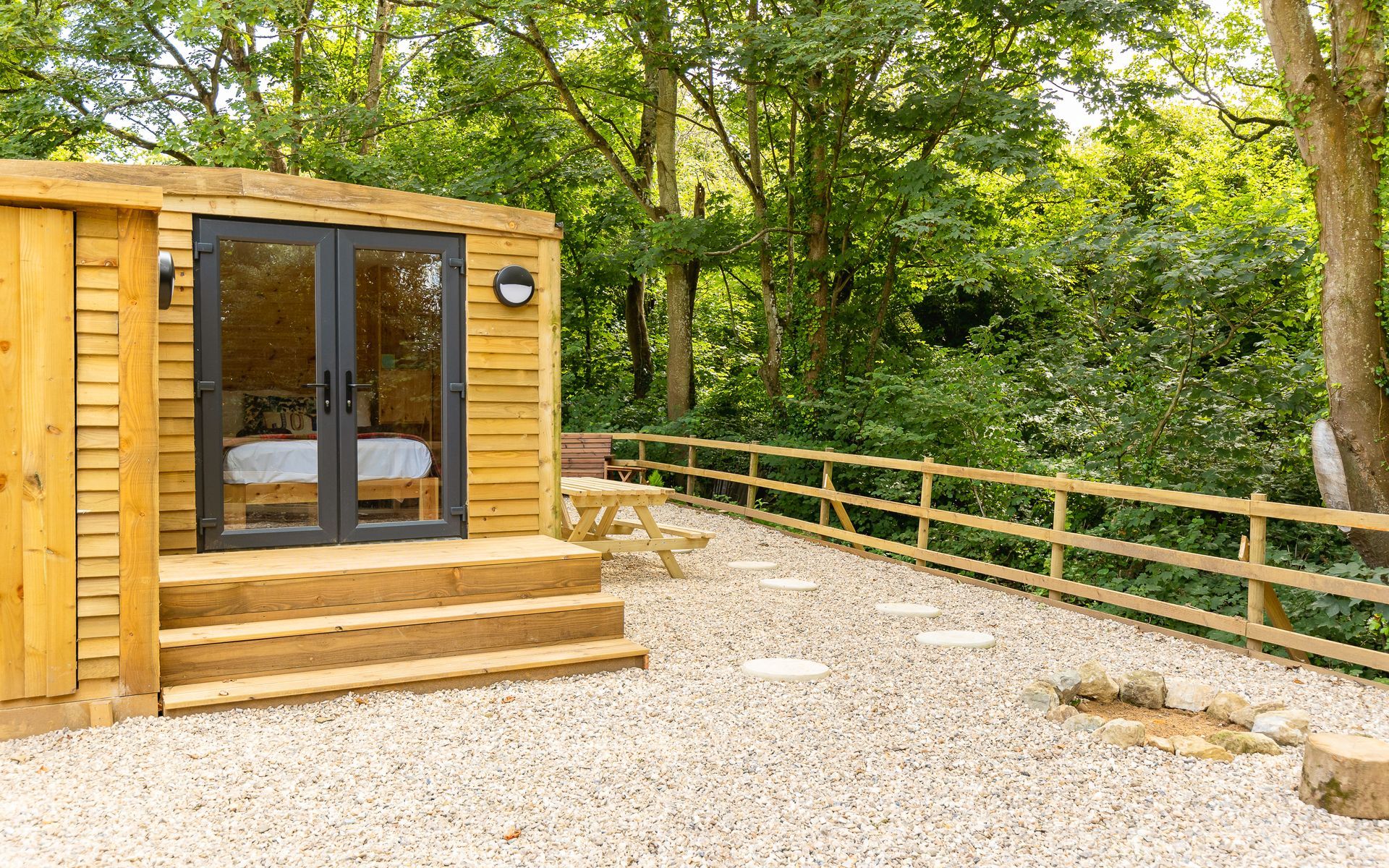 Wooden cabin with steps, double doors, gravel patio, wooden fence, trees.