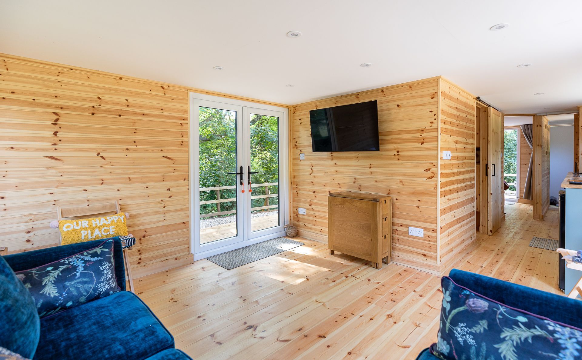 Interior of a cabin with wood-paneled walls and floors, blue armchairs, TV, and glass doors leading outside.