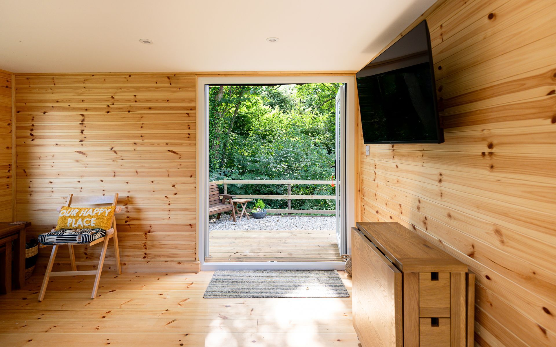 Wooden cabin interior with open door to a deck surrounded by greenery; TV on wall, folding table.