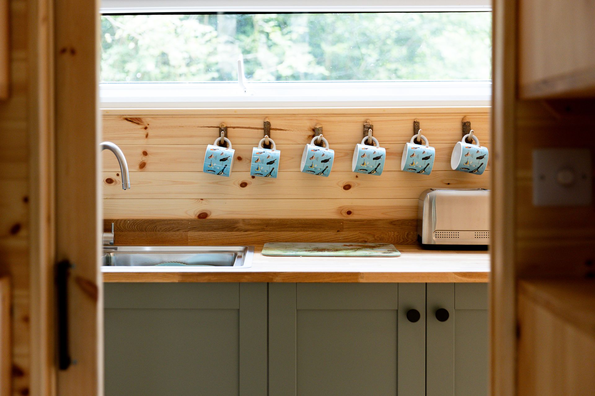 Small cabin kitchen with wooden cabinets, counter, and mugs hanging on hooks.