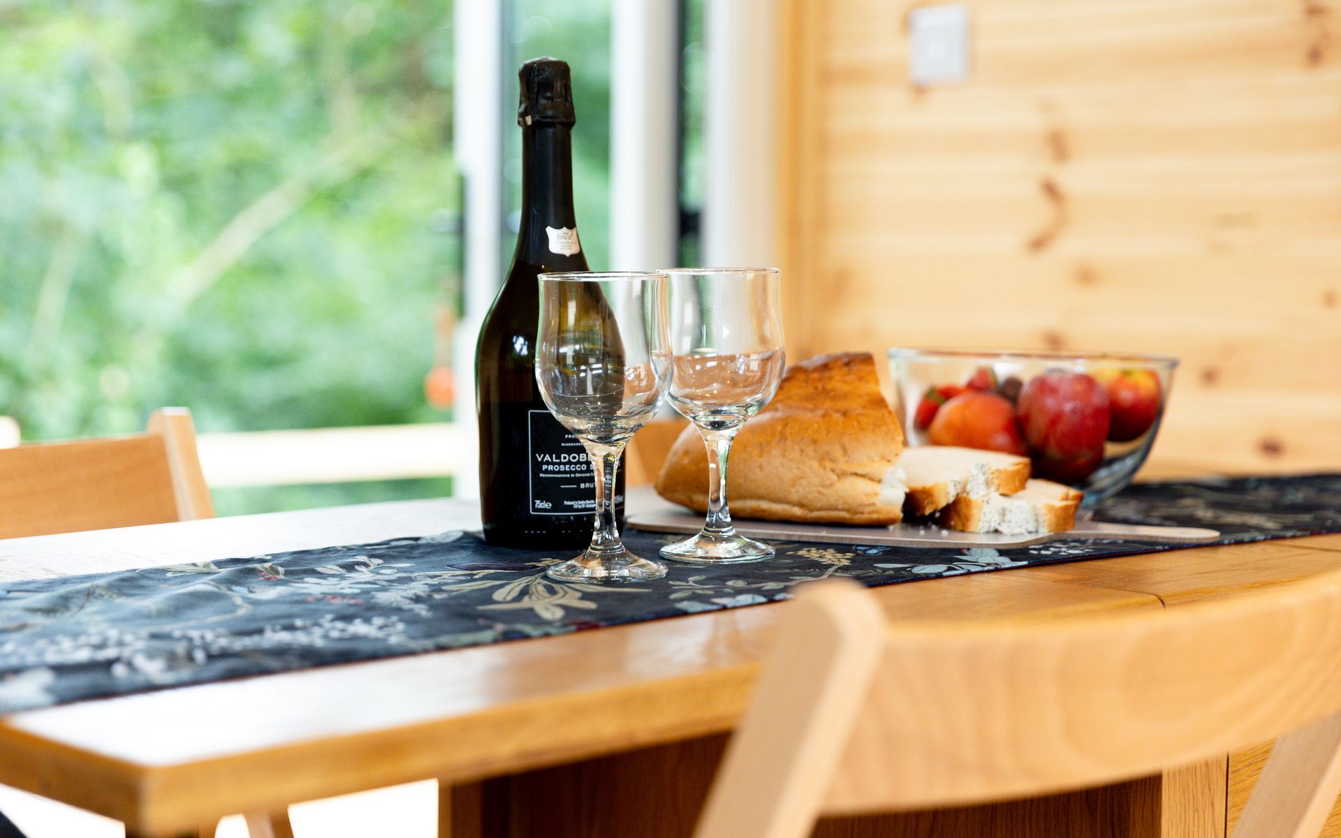 Table set for a meal: wine bottle, glasses, bread, and fruit bowl. Window view in background.