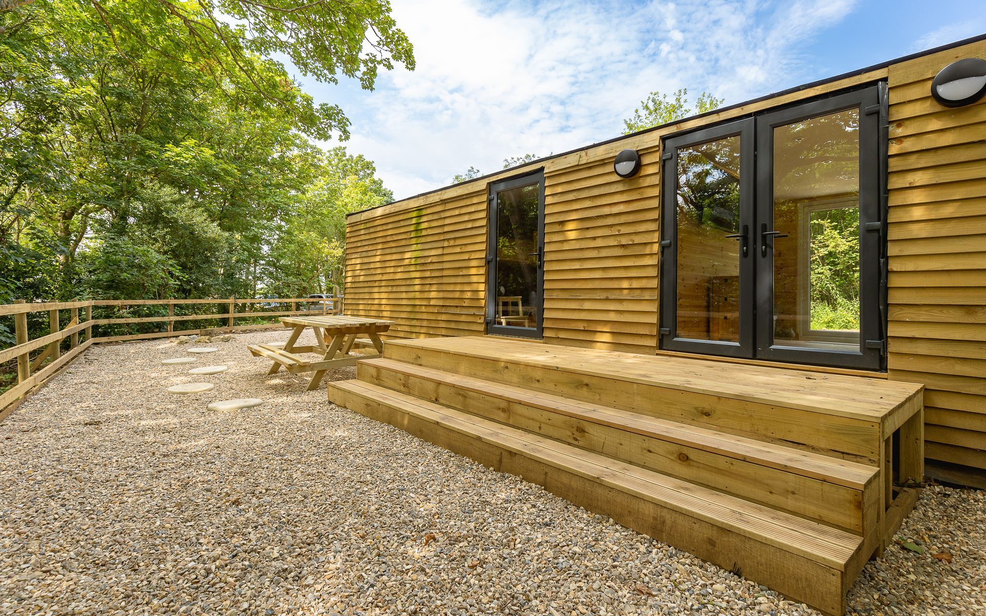 Wooden cabin with deck, picnic table, and French doors; gravel patio, trees in background.
