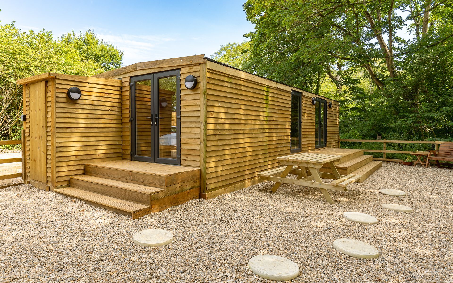 Wooden cabin with steps, a picnic table, and stepping stones in a gravel area surrounded by trees.