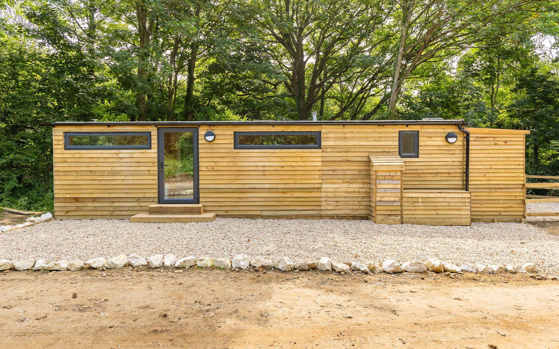 Wooden cabin with glass door, windows, and steps leading up, set on gravel, with trees in background.
