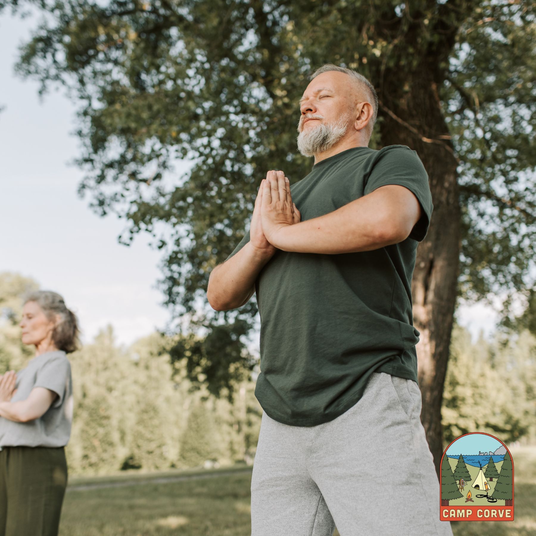 Two people practicing yoga outdoors with hands clasped in front of them, under a tree.