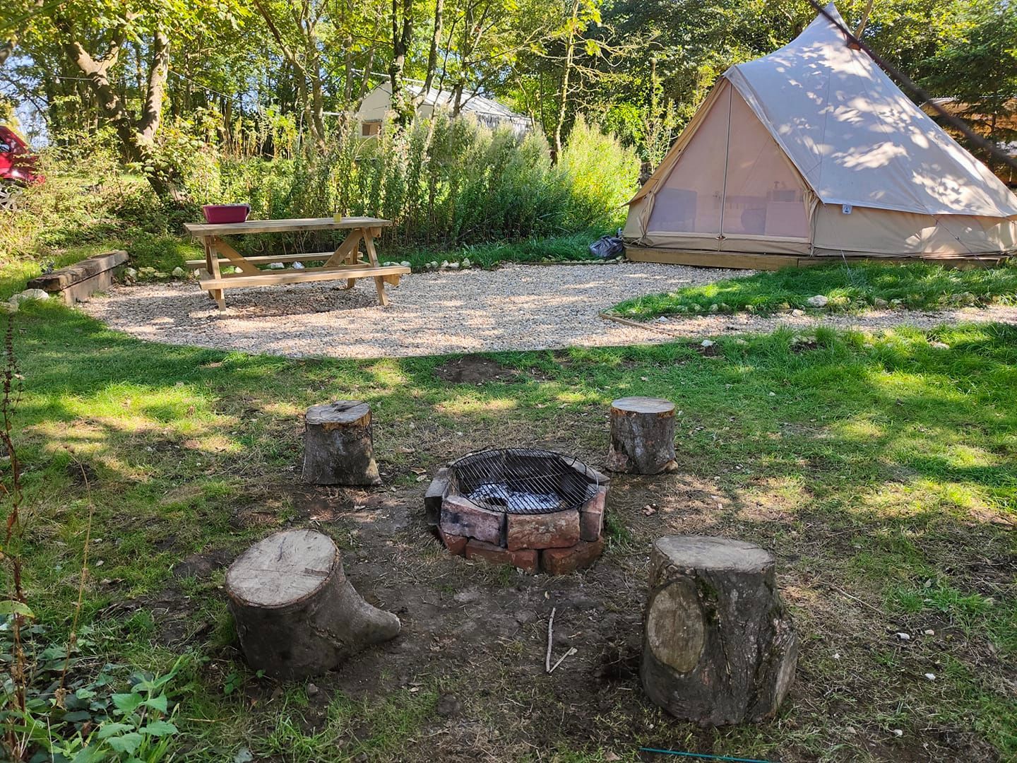 Campground scene: tent, fire pit with logs, picnic table, gravel area, surrounded by trees.
