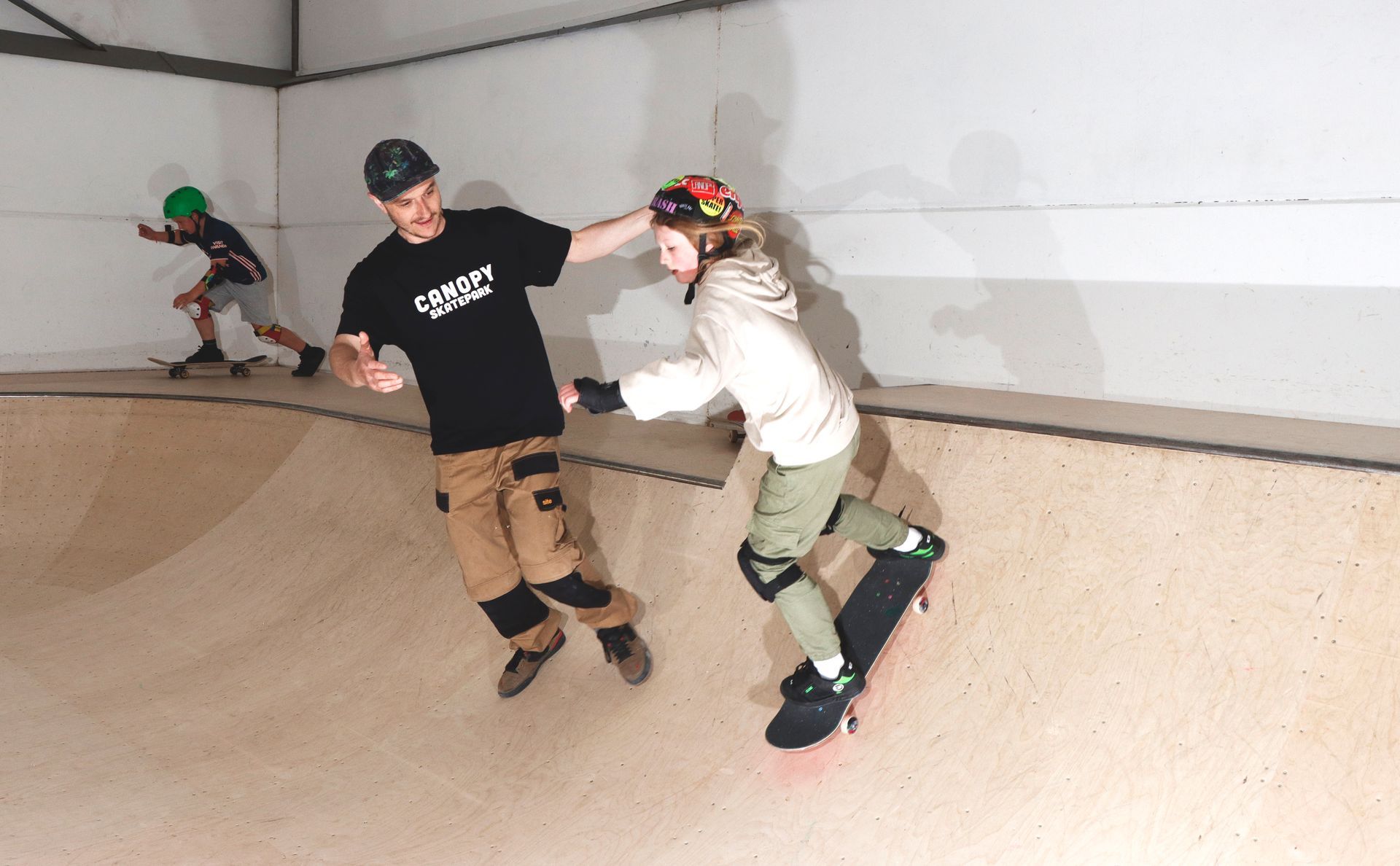 Skateboarder being guided by instructor in a skate bowl. The instructor is supporting the skater. Another skater is in the background.