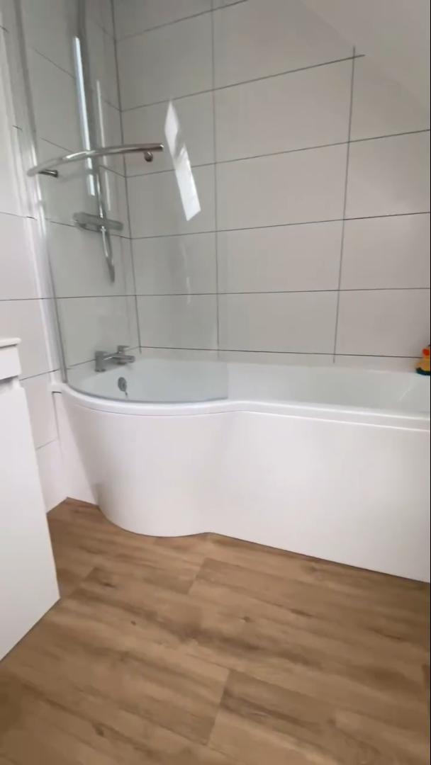 Bathroom with white tub, glass shower screen, and wood-look flooring. White tiled walls.
