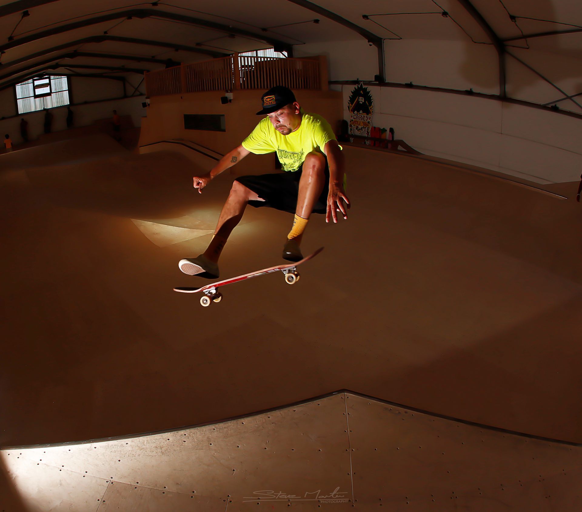 Skateboarder in mid-air above a ramp, indoors. Wearing a yellow shirt and black shorts.