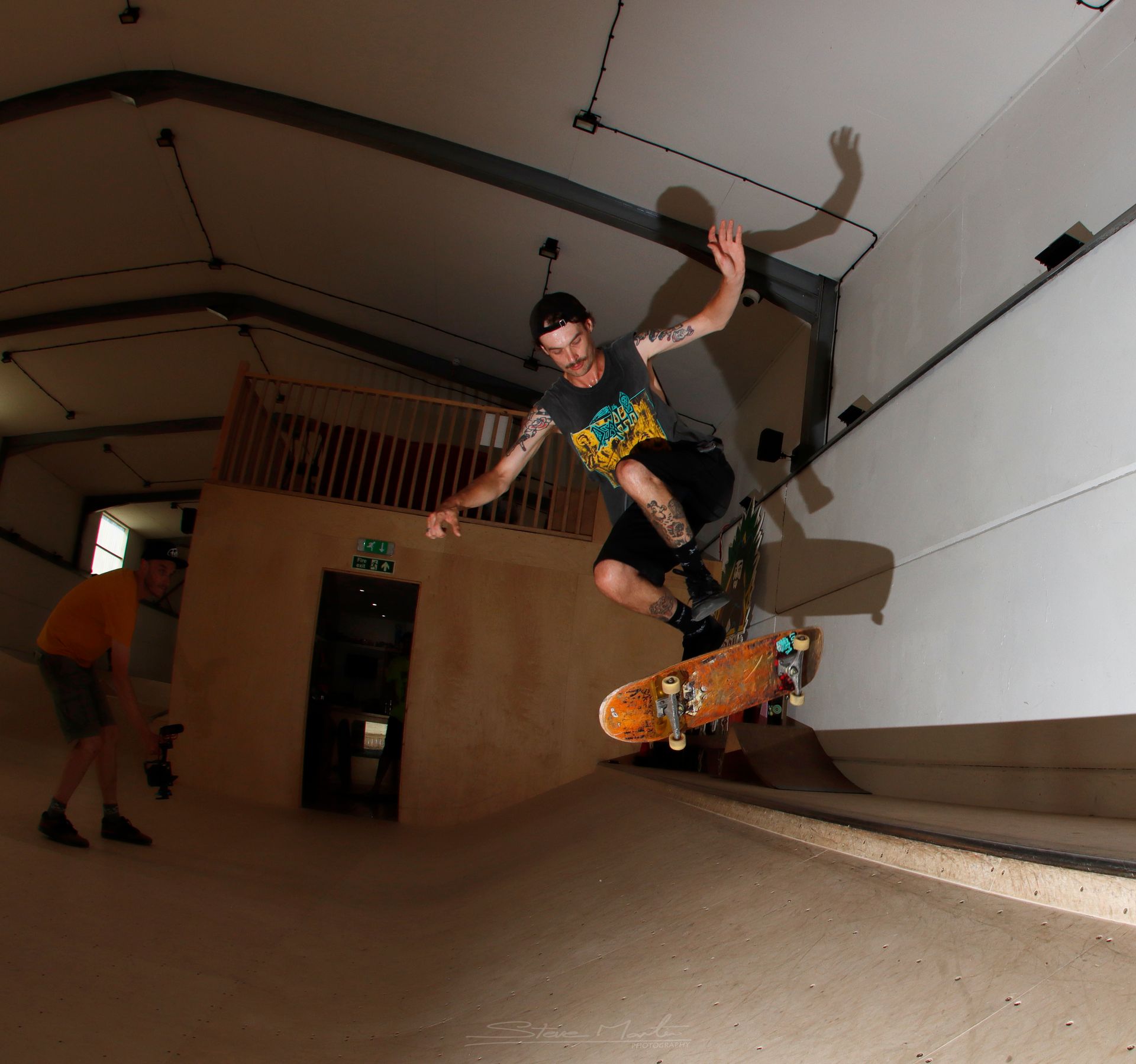 Skateboarder in mid-air on a ramp inside a building. Person with camera stands nearby.