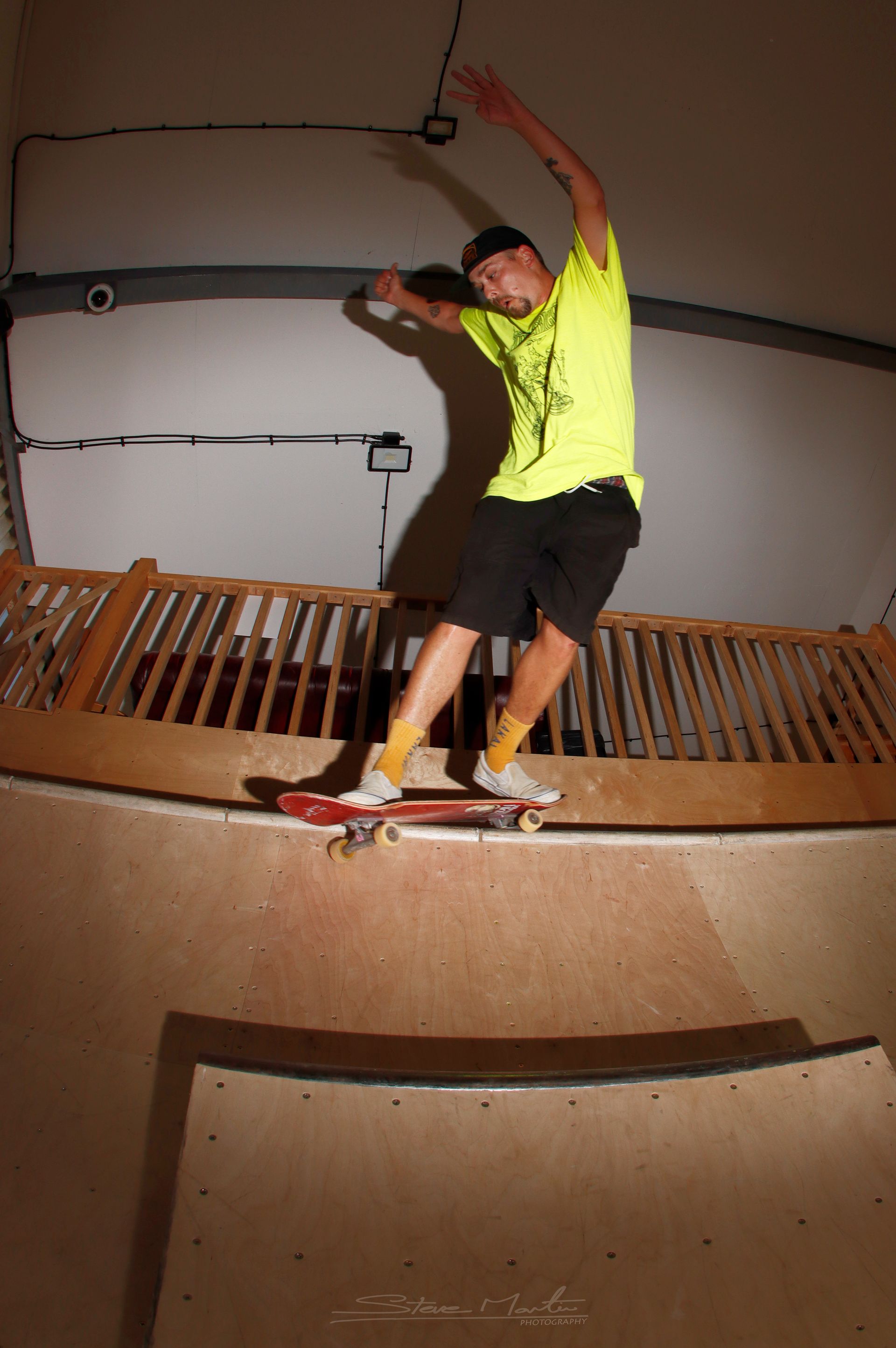 Skateboarder on a ramp, balancing. Yellow shirt, black shorts. Indoor skate park setting.