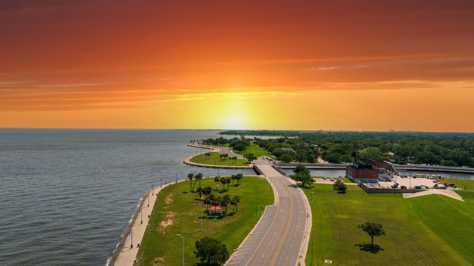 An aerial view of a road leading toward a bridge over water during a vibrant, orange-toned sunset.