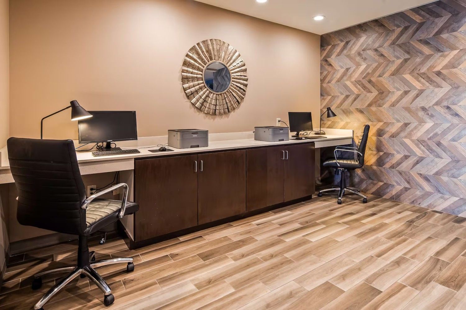 A business center with two computer stations, rolling chairs, and dark wood cabinetry against a patterned wood accent wall.