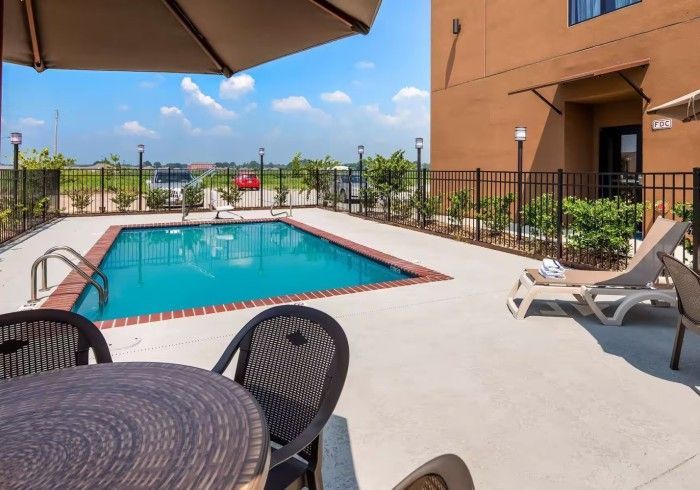 An outdoor swimming pool surrounded by a concrete deck with patio chairs, lounge chairs, and an umbrella, near a building.