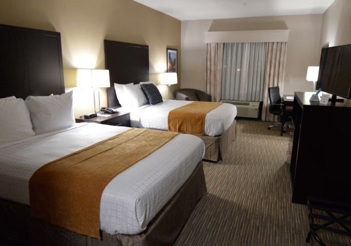 A hotel room with two queen beds featuring white bedding, tan runners, dark headboards, and a desk with a TV.