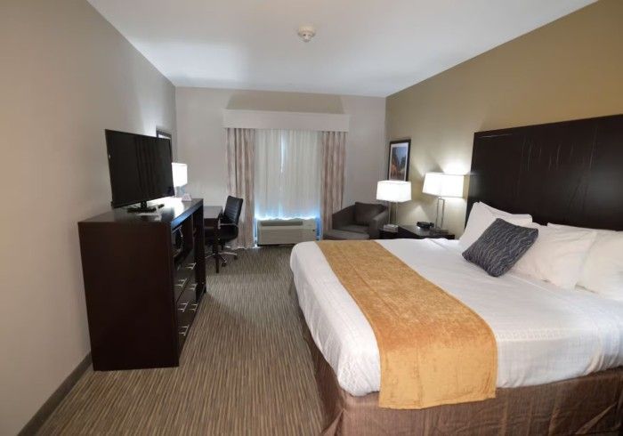 A hotel room featuring a king-size bed with white bedding, a brown runner, a dark wood headboard, and a television dresser.