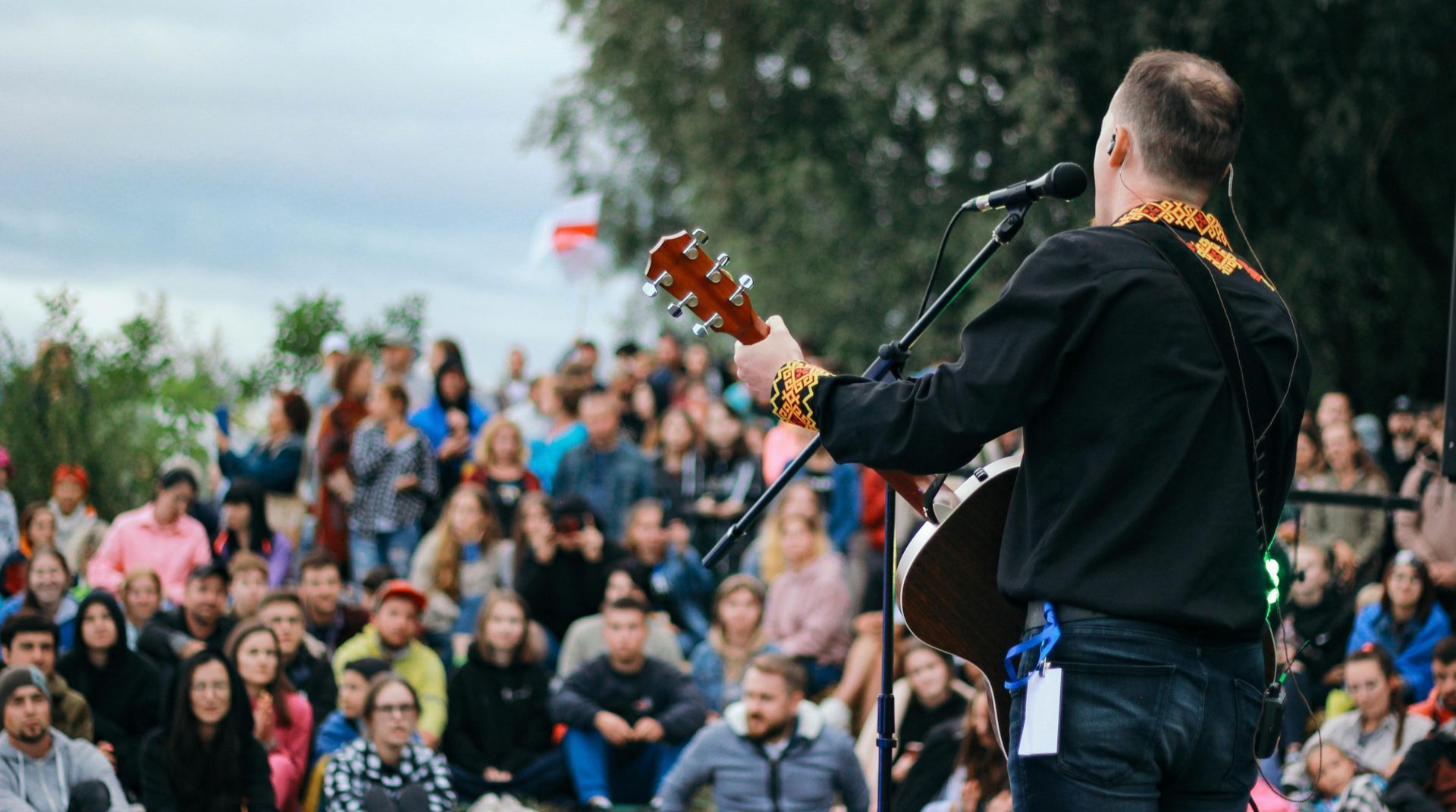 A musician plays an acoustic guitar on stage while performing for a blurred, large outdoor audience.