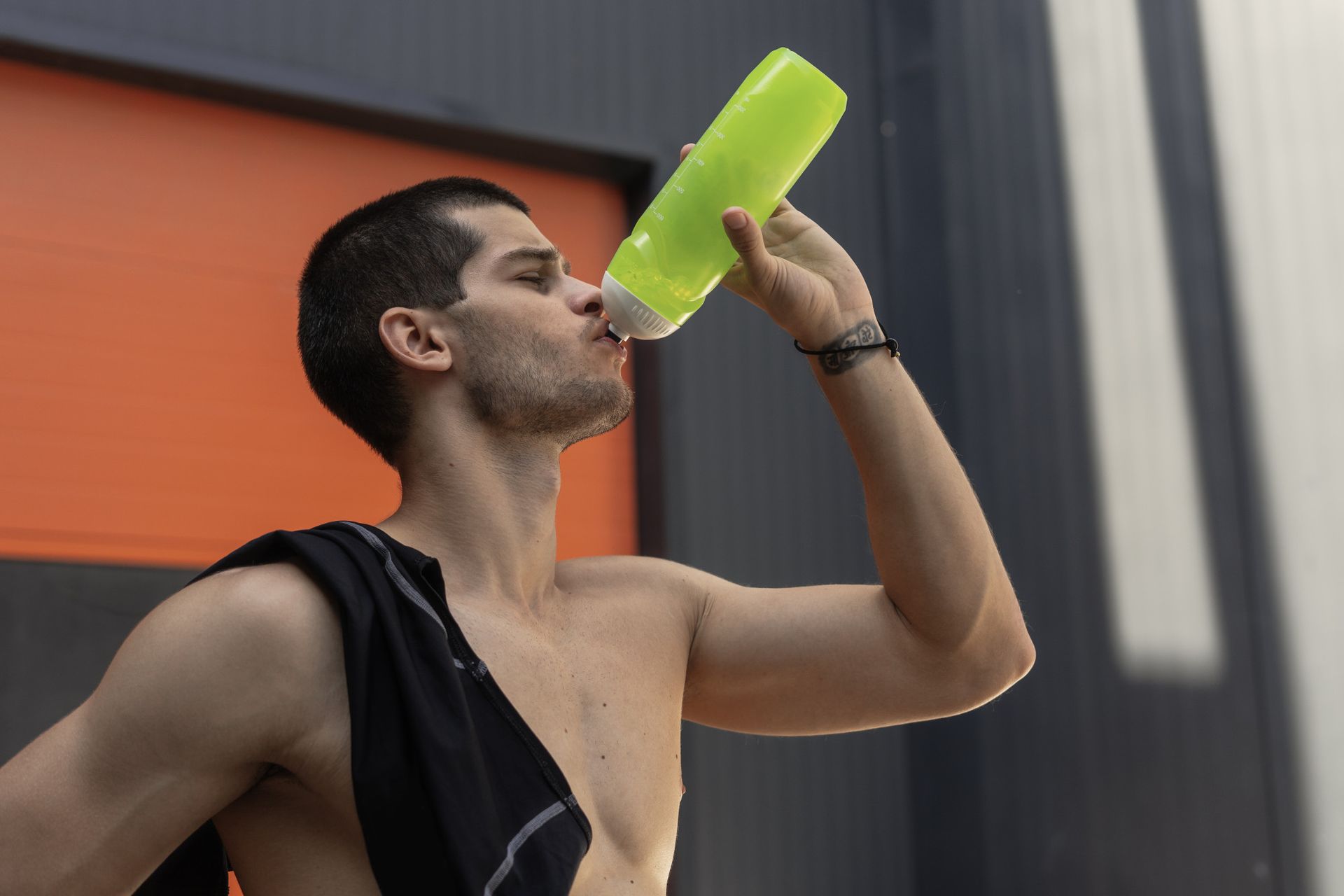 Un atleta sin camisa bebe de una botella de agua de color verde lima con una toalla negra sobre su hombro.